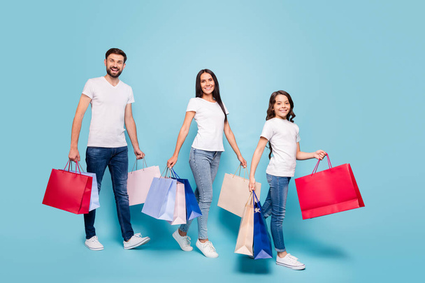 Full length photo of beautiful three people clients mom dad schoolkid with brown hair shop hold bags bargain black friday wear white t-shirt denim jeans sneakers isolated over blue color background - Фото, изображение