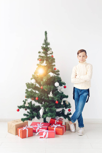 Tatiller ve kış çocukluğu konsepti - gülümseyen genç çocuk beyaz arka planda noel ağacının yanında duruyor - Fotoğraf, Görsel