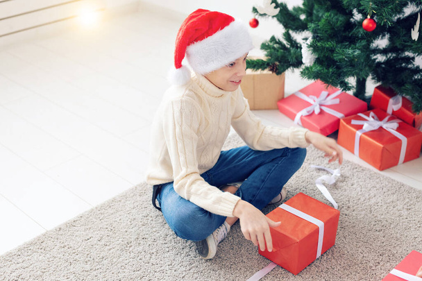 Święta, Boże Narodzenie, dzieciństwo i koncepcja ludzi - uśmiechnięty szczęśliwy chłopak nastolatek w kapeluszu Santa otwiera pudełko na tle choinki - Zdjęcie, obraz