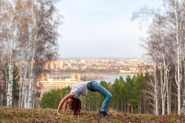 Ένα νεαρό κορίτσι φτιάχνει μια ακροβατική γέφυρα στη φύση με φόντο μια θολή πόλη και δάσος. Το κορίτσι έχει κόκκινα μαλλιά, τζιν και μπλουζάκι. Ακροβατικά στη φύση. Χόρτο στο έδαφος. - Φωτογραφία, εικόνα
