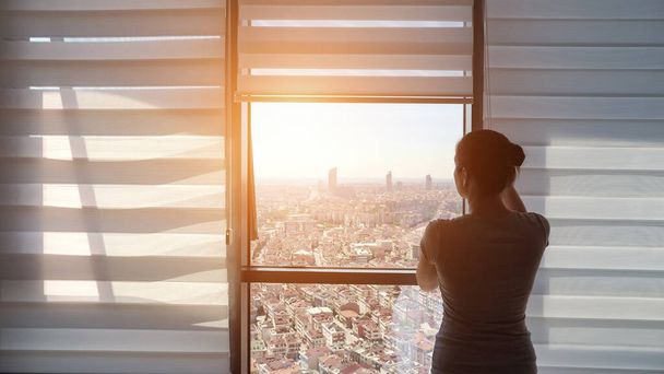 Vrouw opent rolluiken, kijkend naar raam met panoramisch uitzicht op de stad - Foto, afbeelding