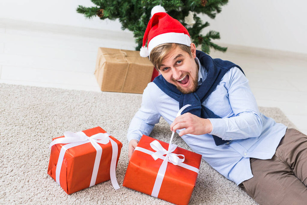 Weihnachts- und Feiertagskonzept - glücklicher Mann mit Weihnachtsmütze öffnet ein Geschenk zu Hause im Wohnzimmer - Foto, Bild
