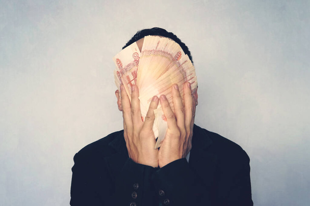 Nuori menestynyt mies pesee kasvonsa rahalla. 5000 seteliä taitettu tuuletin peittää liikemiehen kasvot. lähikuva miehestä, jolla on hallussaan venäläisiä ruplaa
 - Valokuva, kuva