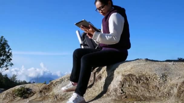 Egy fiatal nő ül a sziklán, és átkutatja a GPS koordinátákat táblagépen, az Inthanon hegy csúcsán, reggel, a nyári vakáció napján. Életmód túra koncepció. - Felvétel, videó