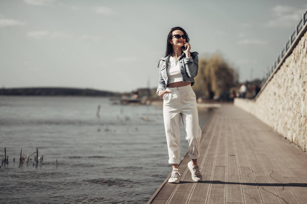Belle fille brune en lunettes de soleil pantalon blanc et veste de jeans près du lac
 - Photo, image
