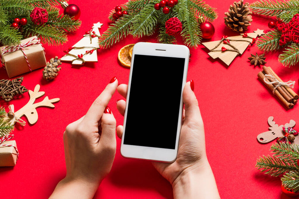 Top view of phone in female hand on festive red background. Christmas decorations. New Year time holiday. Mockup - Photo, Image
