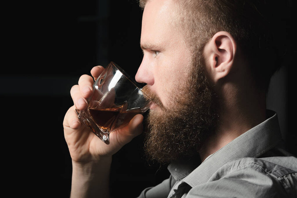 Portrait of a handsome young man with a beard drinking alcohol from a glass. - Фото, изображение
