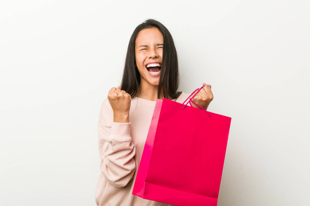 Young hispanic woman holding a shopping bag cheering carefree and excited. Victory concept. - Φωτογραφία, εικόνα
