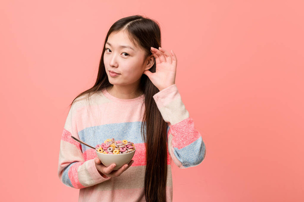 Giovane donna asiatica con una ciotola di cereali cercando di ascoltare un pettegolezzo
. - Foto, immagini
