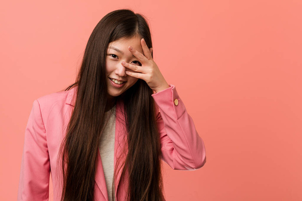 Fiatal üzleti kínai nő rózsaszín öltöny pislogás a kamera ujjain keresztül, zavarban fedő arc. - Fotó, kép