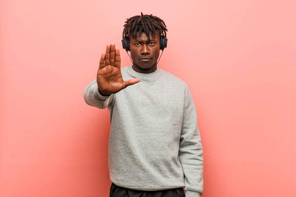Młody rasta czarny człowiek słucha muzyki ze słuchawkami stojącymi wyciągniętą ręką pokazując znak stopu, uniemożliwiając Ci. - Zdjęcie, obraz