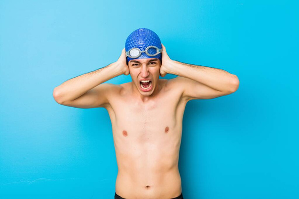 Young swimmer man covering ears with hands trying not to hear too loud sound. - 写真・画像