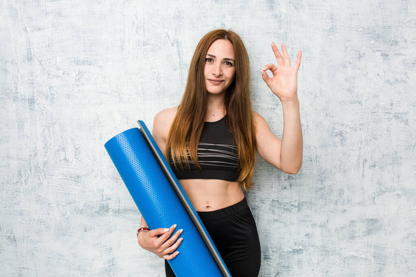 junge kaukasische Sportlerin hält eine Matte fröhlich und zuversichtlich zeigt ok Geste. - Foto, Bild
