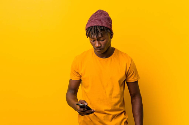 Молодий африканський чоловік стоїть на жовтому тлі в капелюсі і використовує телефон
 - Фото, зображення