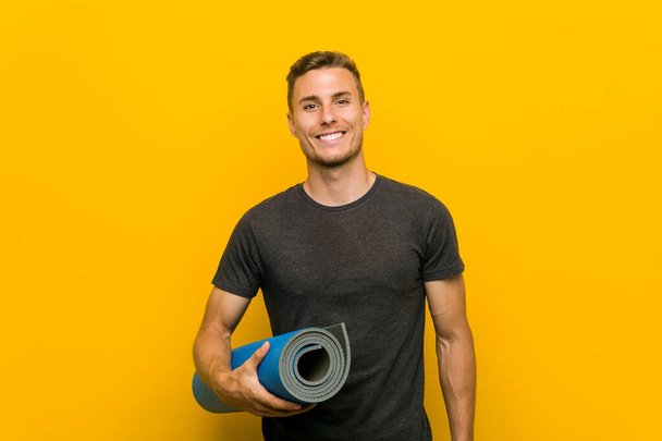 Young caucasian man holding a mat happy, smiling and cheerful. - Photo, Image
