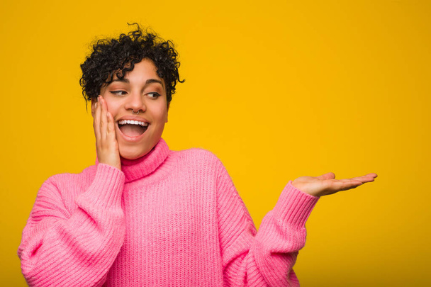 Young african american woman wearing a pink sweater holds copy space on a palm, keep hand over cheek. Amazed and delighted. - Фото, изображение