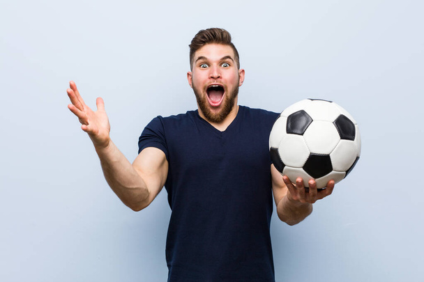 Νέος Καυκάσιος άνθρωπος που κρατά μια μπάλα ποδοσφαίρου γιορτάζοντας μια νίκη ή επιτυχία - Φωτογραφία, εικόνα