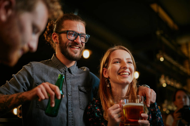 Handsome caucasian man with eyeglasses hugging his girlfriend and talking to her while standing in a pub and drinking beer. Nightlife. - Photo, Image