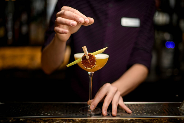 Barman professionnel décorant cocktail alcoolique jaune dans le verre avec un citron séché et feuille tropicale dans la pince à linge
 - Photo, image