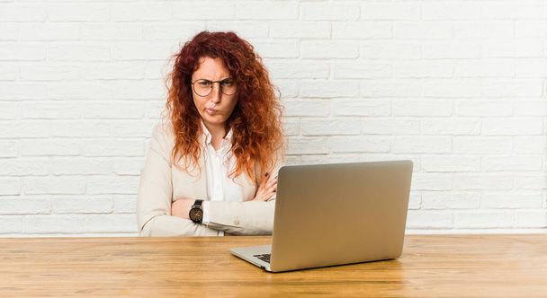 Молода руда кучерява жінка працює зі своїм ноутбуком, що кидається в незадоволення, тримає руки складеними
. - Фото, зображення