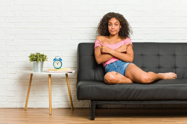 Young african american woman sitting on the sofa points sideways, is trying to choose between two options. - Photo, Image