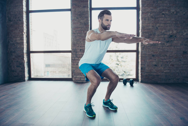 Full length photo of handsome trainer guy doing static squats concentrated sportswear tank-top shorts sneakers training home studio near windows indoors - Foto, Imagem
