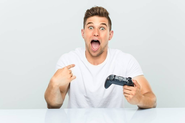 Giovane uomo in possesso di un controller di gioco sorpreso puntando a se stesso, sorridendo ampiamente
. - Foto, immagini