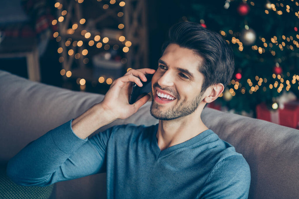 Close up photo of joyful guy speaking cellphone tell congratulations with newyear enjoy christmas atmosphere sit couch in house with bright magic miracle lights - Foto, afbeelding