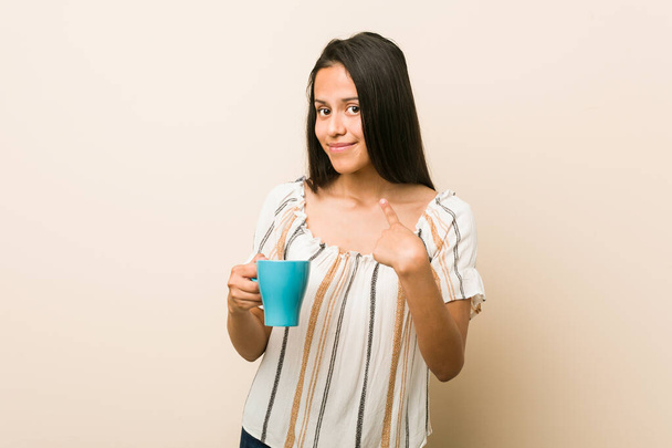 Jovem hispânica segurando uma xícara apontando com o dedo para você como se convidando chegar mais perto
. - Foto, Imagem