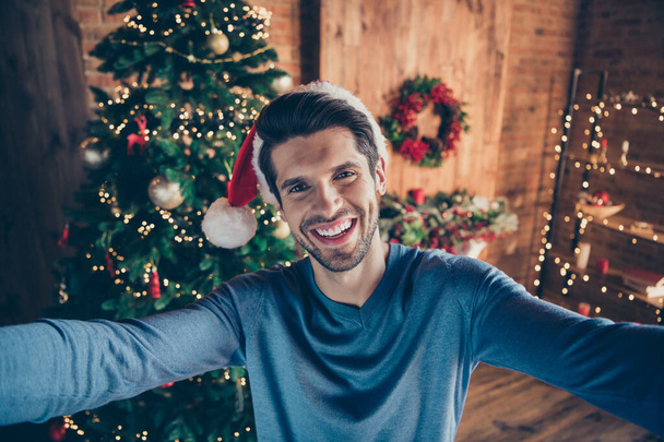 Self portrait of cheerful excited enjoying man having finished decorating his christmas tree and taking selfie on background of it smiling toothily in santa cap - Фото, изображение