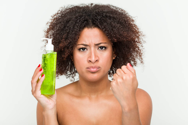 Genç Afrikalı Amerikalı kadın aloe vera şişesi tutuyor kameraya yumruğunu gösteriyor, agresif yüz ifadesi.. - Fotoğraf, Görsel