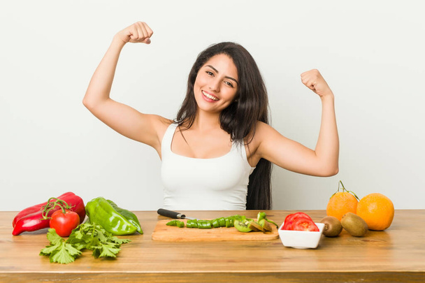Młoda zakrzywiona kobieta przygotowująca zdrowy posiłek pokazujący gest siły ramionami, symbol kobiecej siły - Zdjęcie, obraz