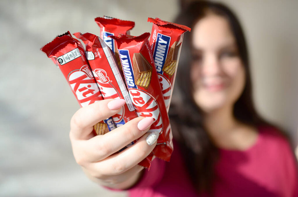 KHARKOV, UKRAINE - OCTOBER 21, 2019: A young caucasian brunette girl shows many kit kat chocolate bars in red wrapping in light room. Kit Kat chocolate manufactured by Nestle - Photo, image