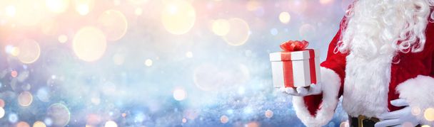 Santa Claus Holding A Christmas Present With Snowy Background And Golden Lights - Photo, image