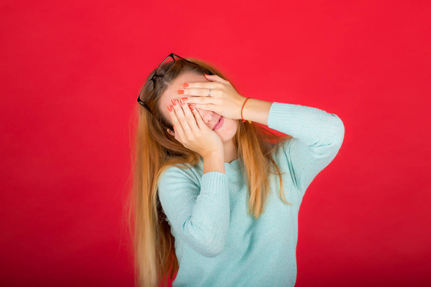 red-haired girl covered her face with her hands, photo studio isolated on red background - Fotó, kép