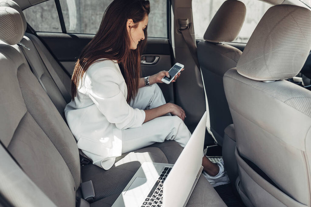 Μόδα κομψό κορίτσι σε λευκό κοστούμι δουλεύει στο φορητό υπολογιστή στο αυτοκίνητο - Φωτογραφία, εικόνα