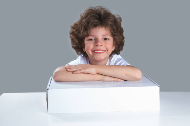 A little boy sits at the table folded his hands in a white box. Smiling looking at the camera. Gray background. Close-up. - Foto, Imagem