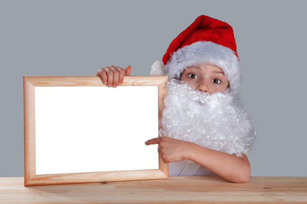 Young Santa holding a blank picture frame and displays it on your finger. He sits at a table looking at the camera. Close-up. Gray background. - Foto, Imagem