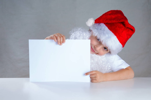 Little Santa holding a white envelope, smiling. Sitting behind a white table looking at the camera. Close-up. Gray background. - Foto, Bild