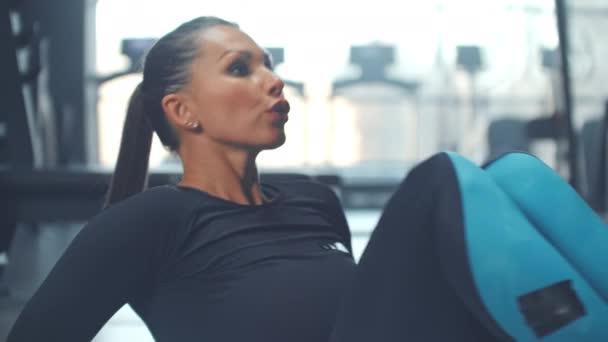 Wysoce zmotywowana kobieta trenuje mięśnie brzucha w siłowni z dużymi oknami na tle biegającego mężczyzny na bieżni - Materiał filmowy, wideo