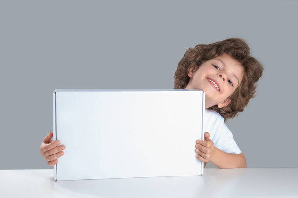 Cute curly little boy sitting at a table holding a white box. He looks into the camera. Gray background. Close-up. - Foto, Imagem