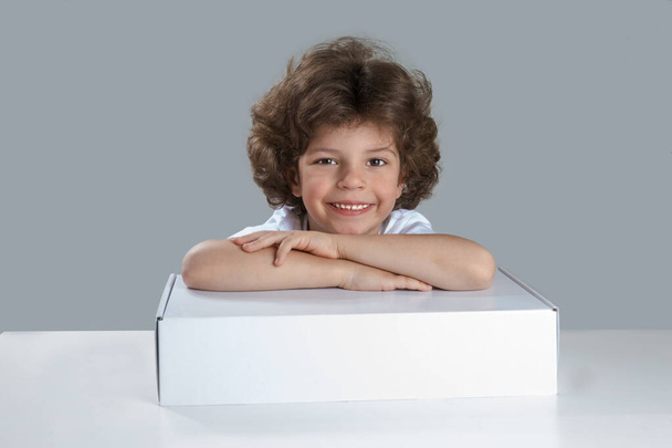Little boy smiling looking at the camera. Sitting at the table folded hands on the white box. Sad looking at the camera. Gray background. Close-up. - Фото, изображение