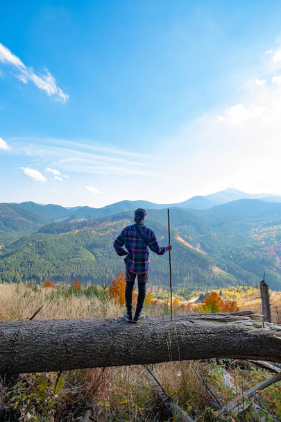 Young male traveler with stick in hands standing on a big log, mountains and sky on background - Foto, afbeelding
