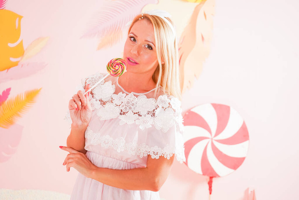 charming happy blonde woman in a soft lace summer dress posing with candy lollipop in the pool with foam soft small balls and engaged in relaxation therapy. - Photo, Image