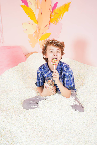 tiener jongen in het zwembad met schuim zachte kleine witte ballen. persoon die zich bezighoudt met ontspanningstherapie. blank kind met kleurrijke lolly op de stok op roze achtergrond. - Foto, afbeelding