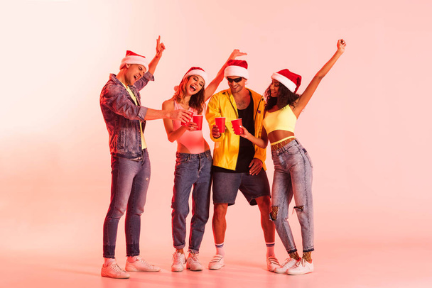 excited men and multicultural girls in santa hats holding plastic cups on pink  - Foto, Bild