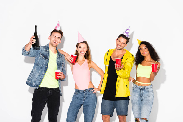 счастливых мультикультурных друзей в шапочках для вечеринок, держащих пластиковые чашки на белом
 - Фото, изображение