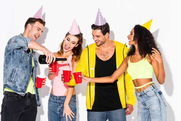 happy man pouring champagne in plastic cups near multicultural friends in party caps on white - Photo, Image