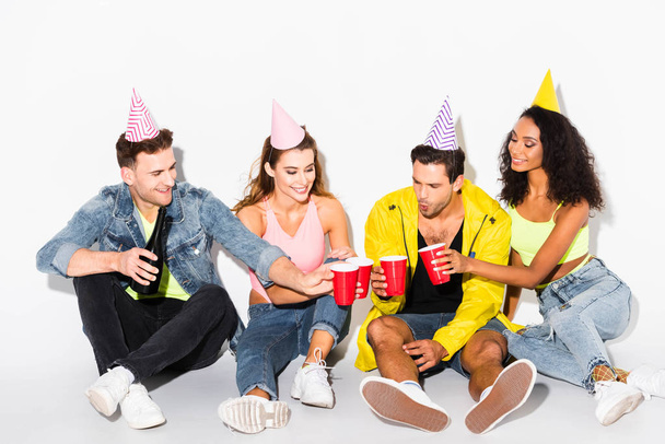 happy man holding plastic cups and clinking with multicultural friends in party caps on white - Foto, immagini