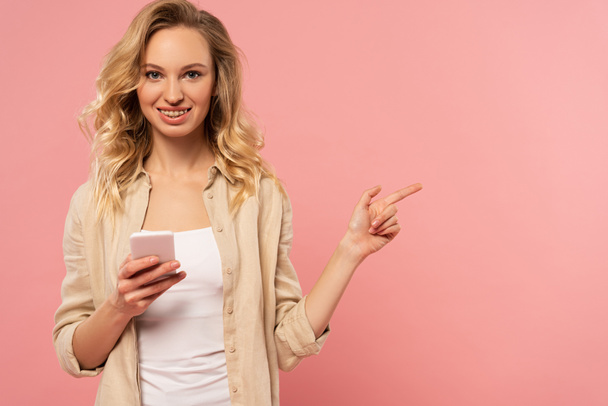 Smiling woman holding smartphone and pointing with finger away isolated on pink - 写真・画像